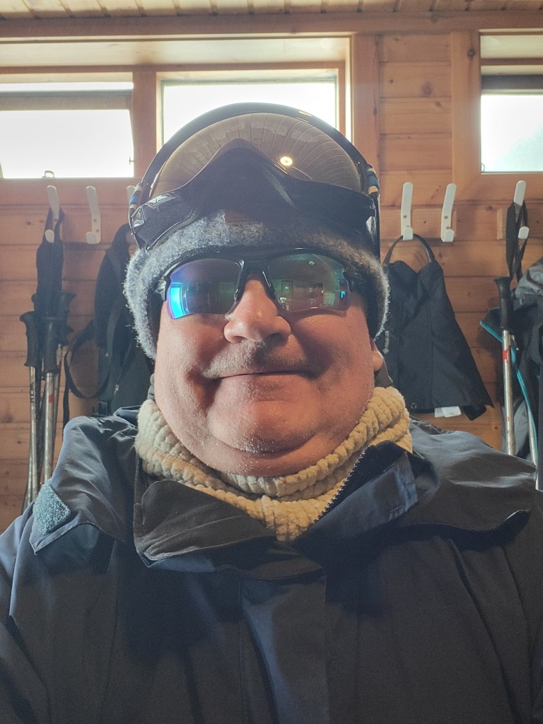 Man standing in ski gear with sunglasses, wooly hat and goggles on his head.